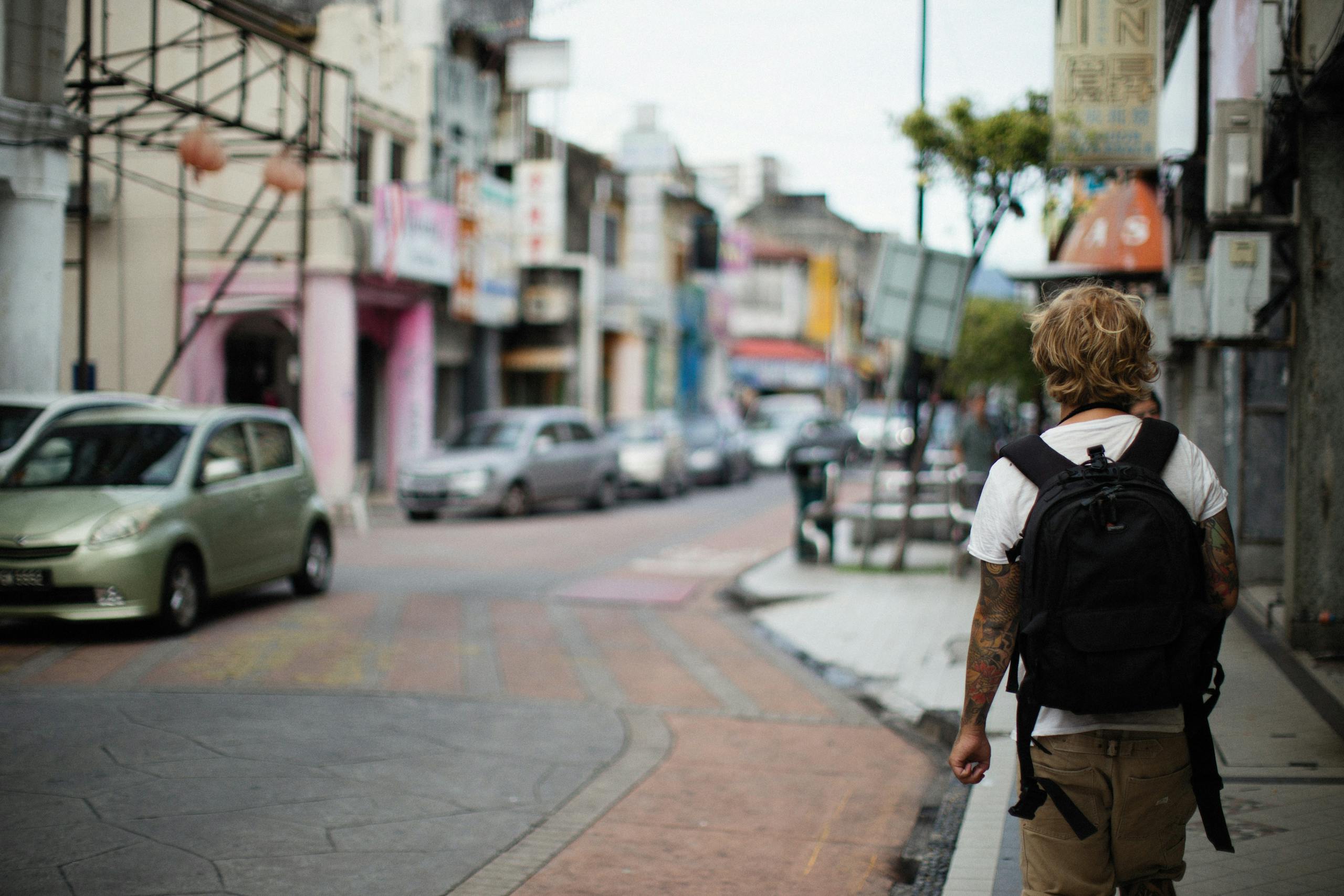 Blonde man with backpack walking on lively city street, captured from behind.