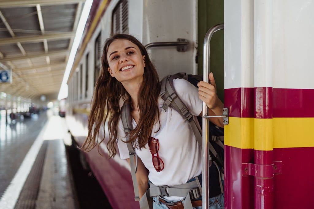 A cheerful woman with a backpack smiling while leaning out of a train at a station.