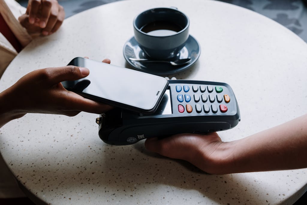 A person using a smartphone to make a contactless payment with a card reader on a cafe table.