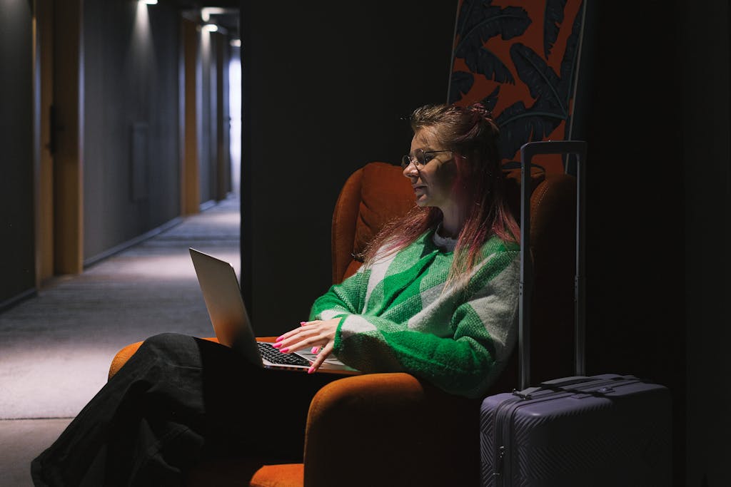 A woman in a hotel hallway using a laptop, sitting on an armchair next to luggage, creating a cozy working environment.