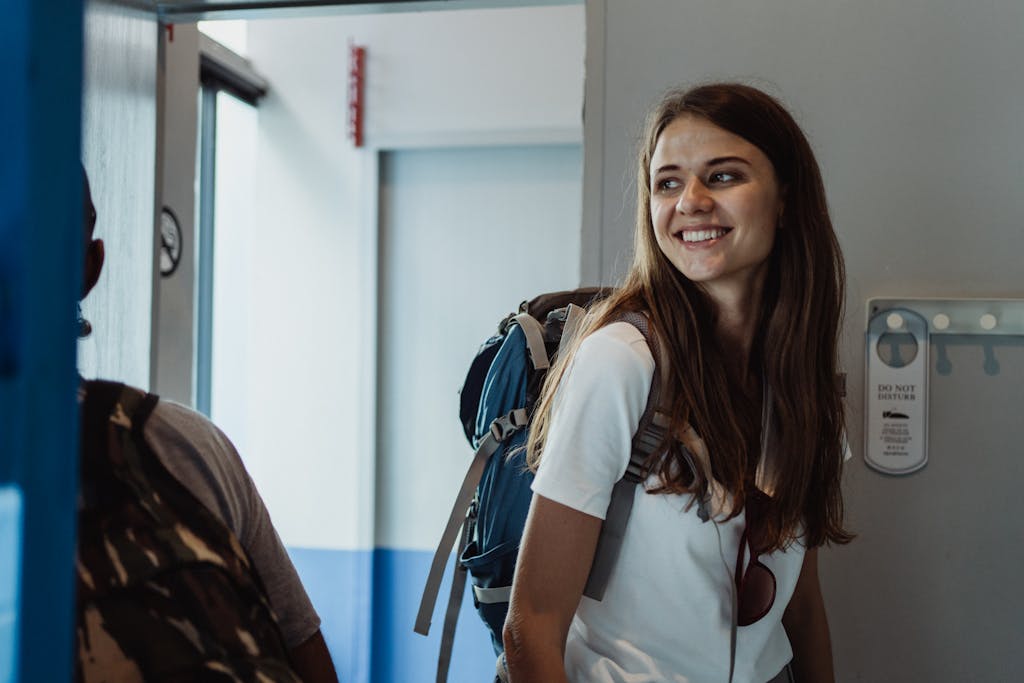 A young woman with a backpack smiles in a hostel hallway, ready for adventure.