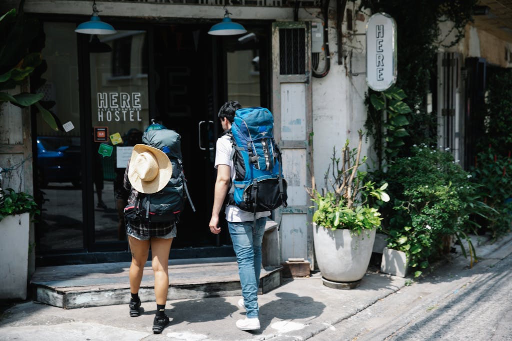 Back view of anonymous couple of tourists with rucksacks walking on street and entering hostel door during trip