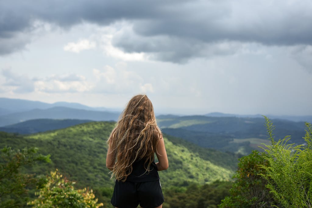 A woman with long hair stands outdoors, overlooking a vast mountainous landscape under a cloudy sky.