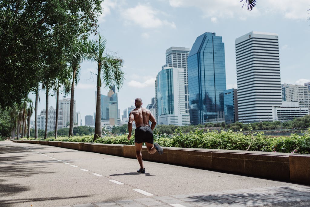 Shirtless man jogging along a city walkway with skyscrapers in the background, emphasizing fitness in an urban setting.